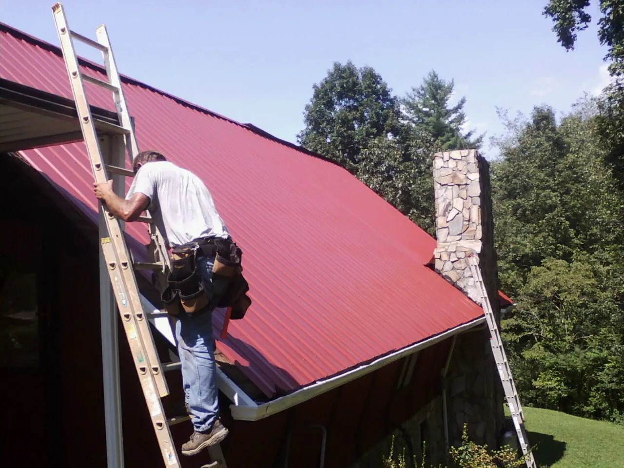 Roofer on a ladder beside a red metal roof and stone chimney — Soffit & Fascia Repair by Roofing Brothers in Tellico Plains